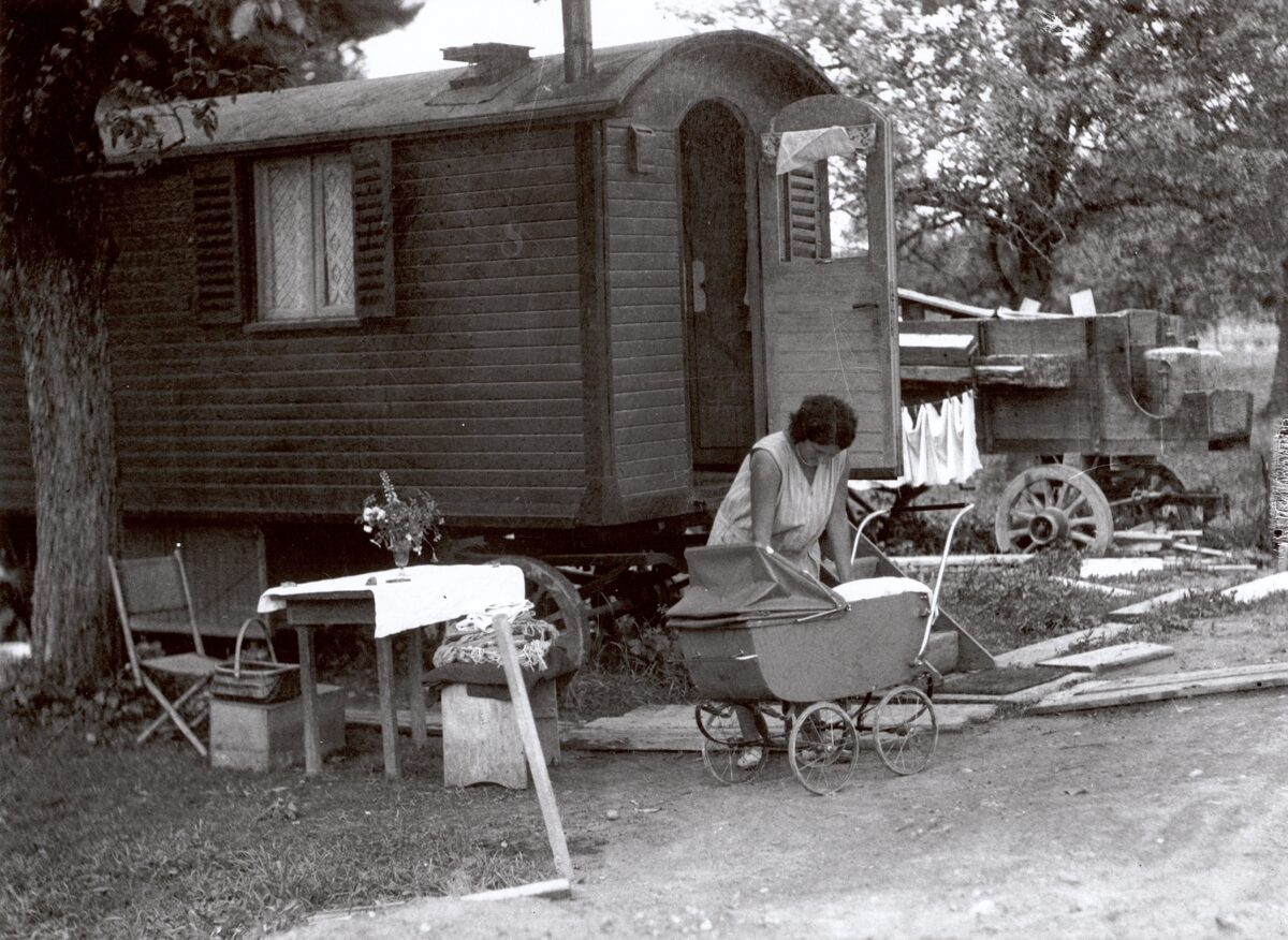 Photographie en noir et blanc d'une femme avec un enfant devant une caravane. L'enfant est dans une poussette. À l'arrière-plan, du linge sèche, au premier plan se trouve une petite table avec un bouquet de fleurs.