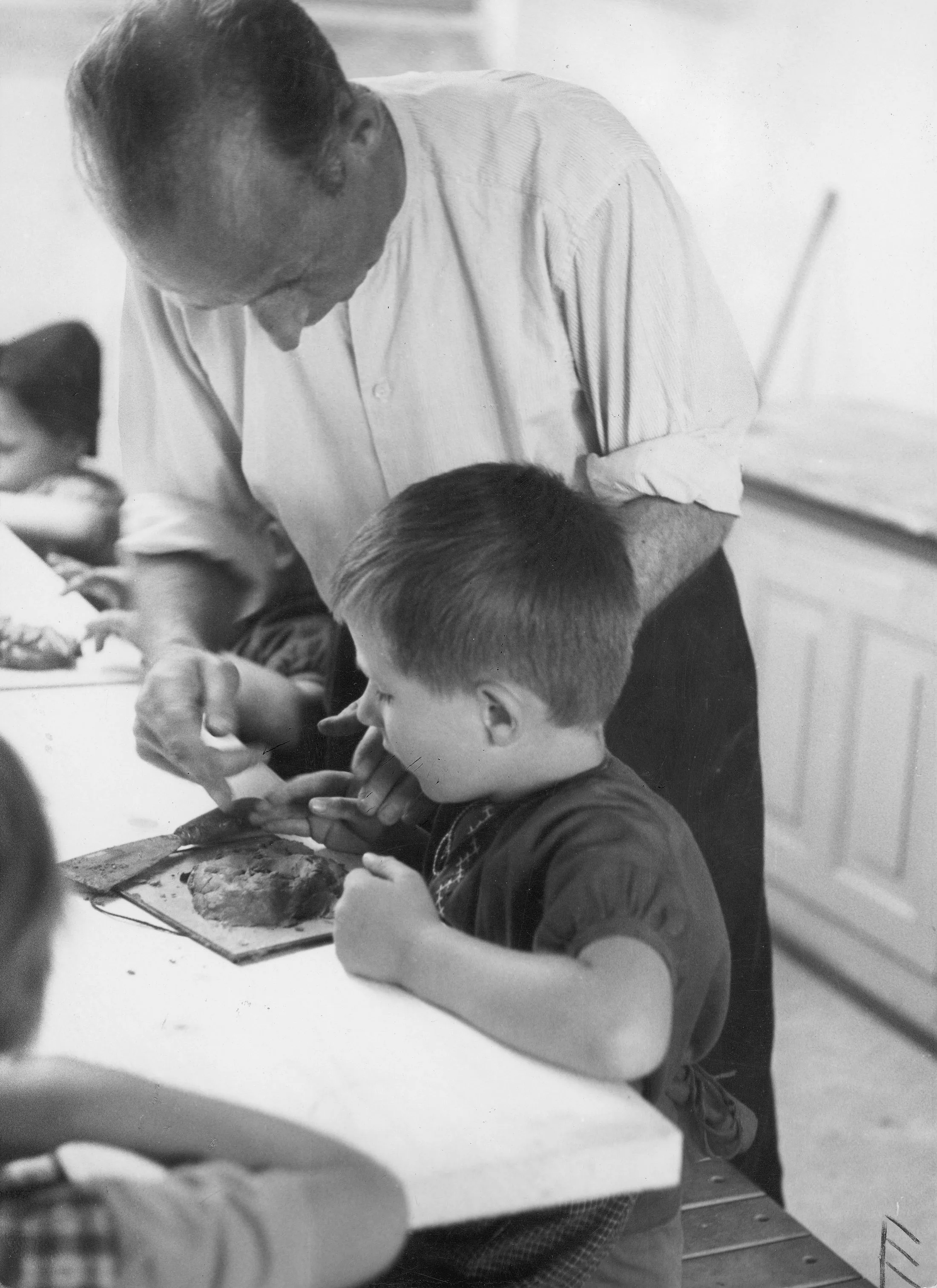 Photographie en noir et blanc d'un homme adulte et d'enfants autour d'une table. L'homme est debout, les enfants sont assis à table et bricolent. Il s'agit des enfants placés et du père adoptif d'une grande famille d'accueil vers 1950.