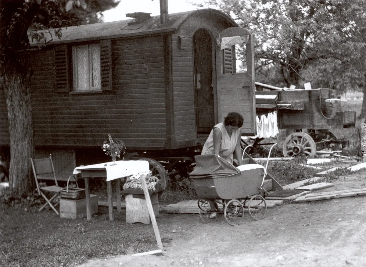 Photographie en noir et blanc d'une femme avec un enfant devant une caravane. L'enfant est dans une poussette. À l'arrière-plan, du linge sèche, au premier plan se trouve une petite table avec un bouquet de fleurs.