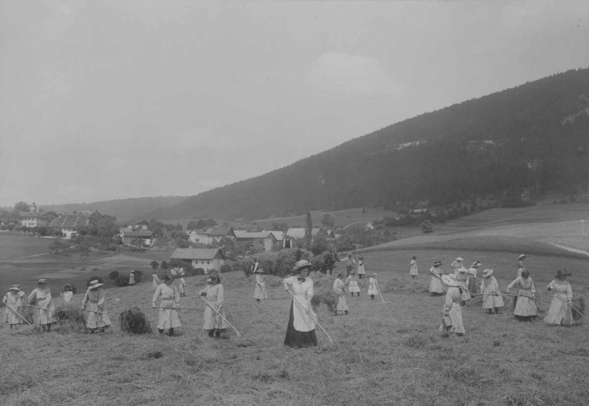 Photographie en noir et blanc de jeunes femmes en habits du dimanche travaillant dans les champs à la maison d'éducation de Loveresse. La photo provient du volume 2 des albums photos de la Direction de l'aide aux indigents du canton de Berne et a été exposée à l'Exposition nationale de 1914 à Berne.